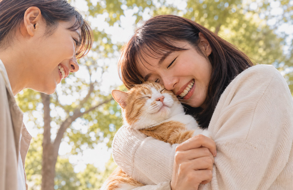 “在溫暖陽光下，兩位幸福的亞洲寵物飼主在戶外親密擁抱一隻可愛的橘白貓，表情充滿愛與幸福。這張圖片展示了寵物與飼主之間強烈的情感紐帶，與 ICE 寵物緊急聯絡卡的主題相呼應。”
